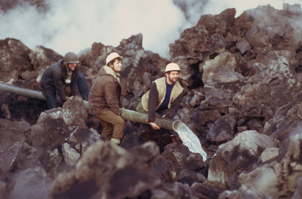Three white men wearing helmets hold a pipe pouring water out in a rocky, steamy landscape.