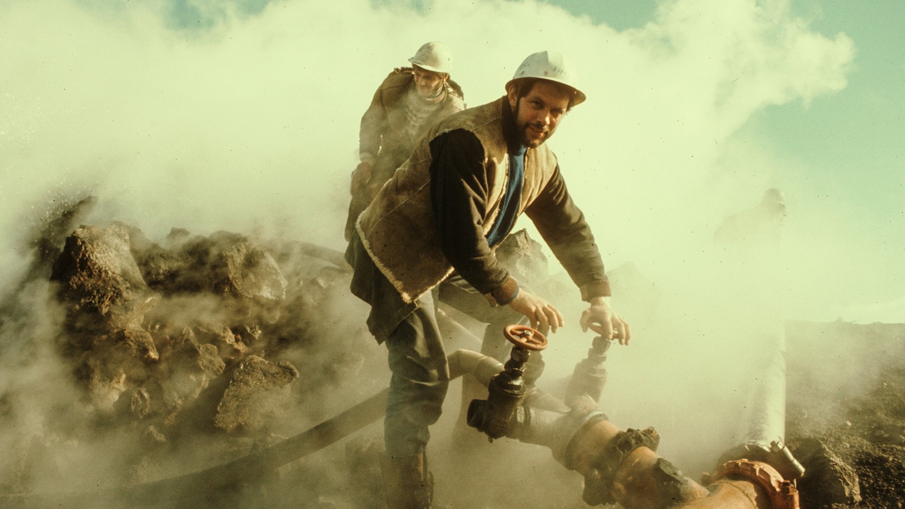 Two men in a steamy landscape with a pipe. One man has his hands on two valves.