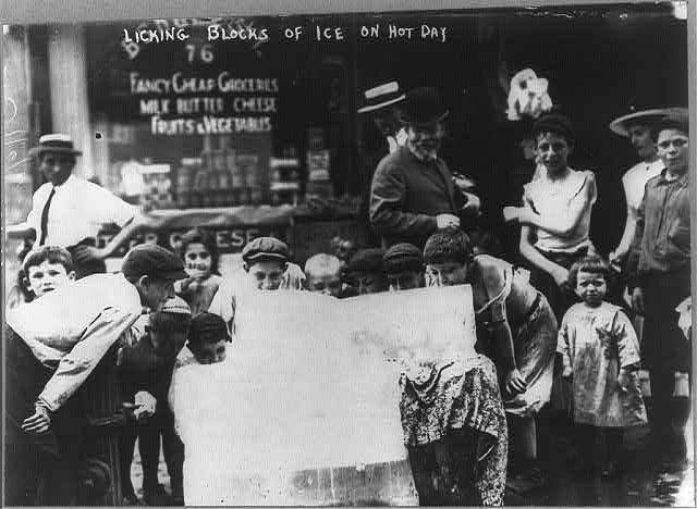 Photograph shows a group of children licking large blocks of ice on the sidewalk in front of a grocery store. 