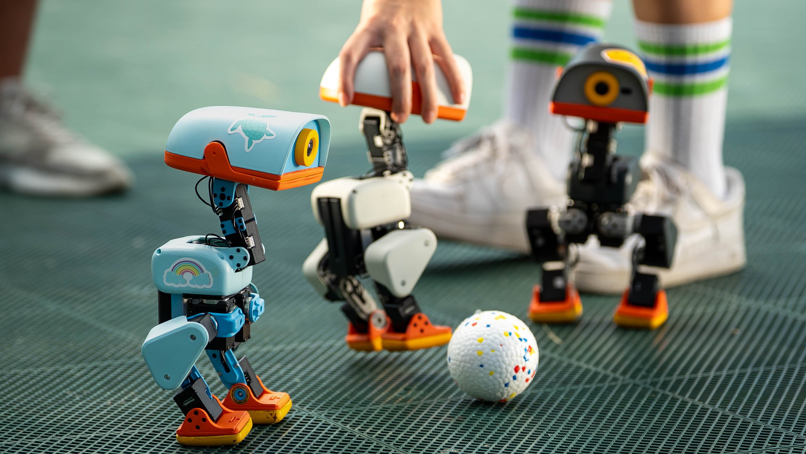 three small robots with two legs and one eye stand around a white ball with yellow, blue, and red flecks on it