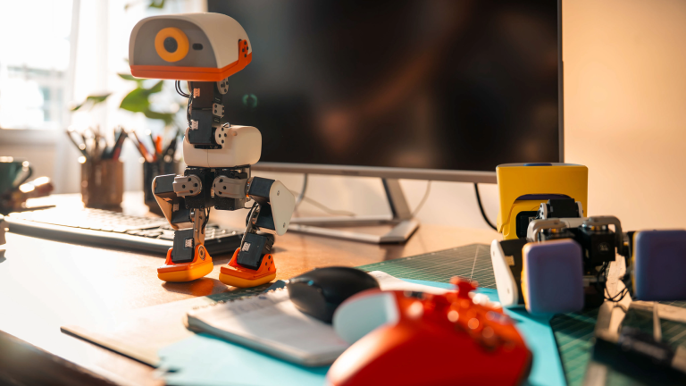 a robot with two legs and one eye stands on a desk with a computer screen