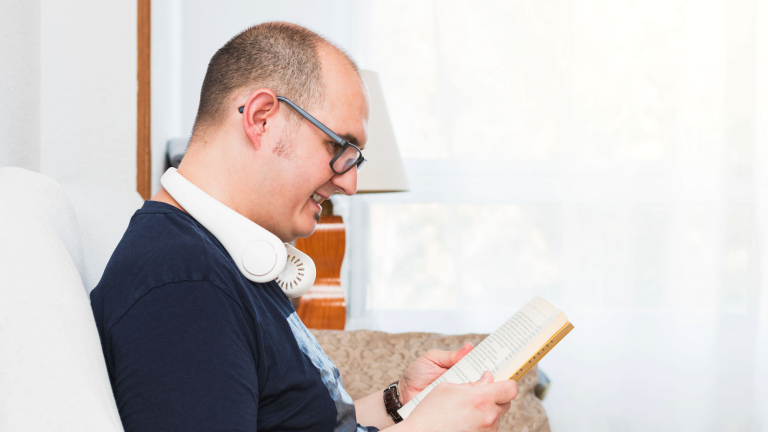 a man sitting on a couch reading a book and wearing a neck fan