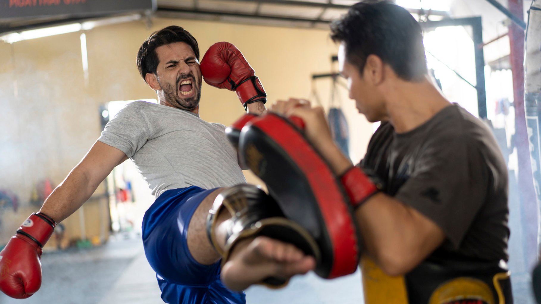 A man with dark hair, white shirt, and blue shorts wearing red boxing gloves kicks a pad another asian man is holding in a garage workout space.