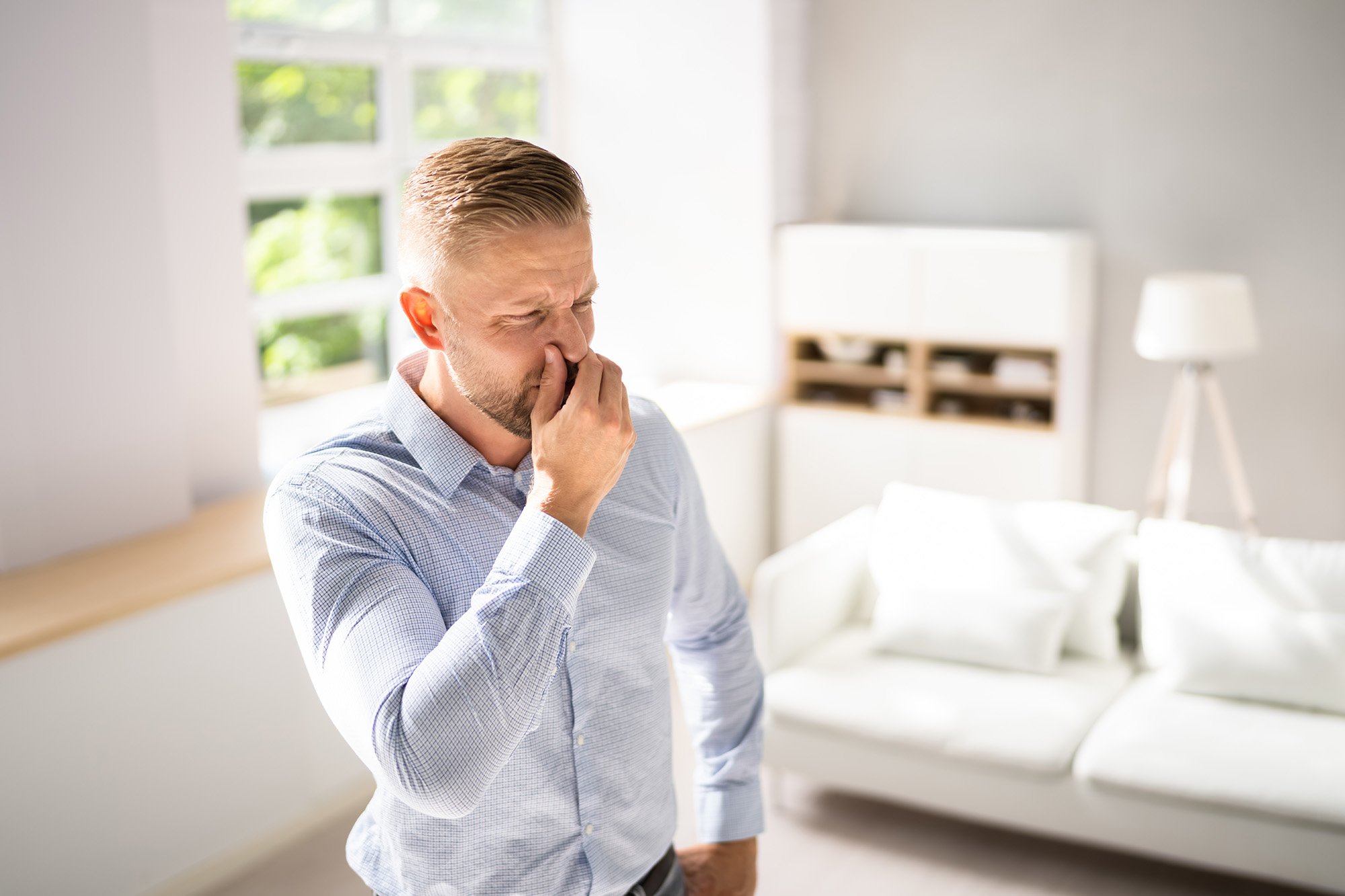 A man standing in a living room holding his nose and mouth like he smells a stink or he's about to sneeze