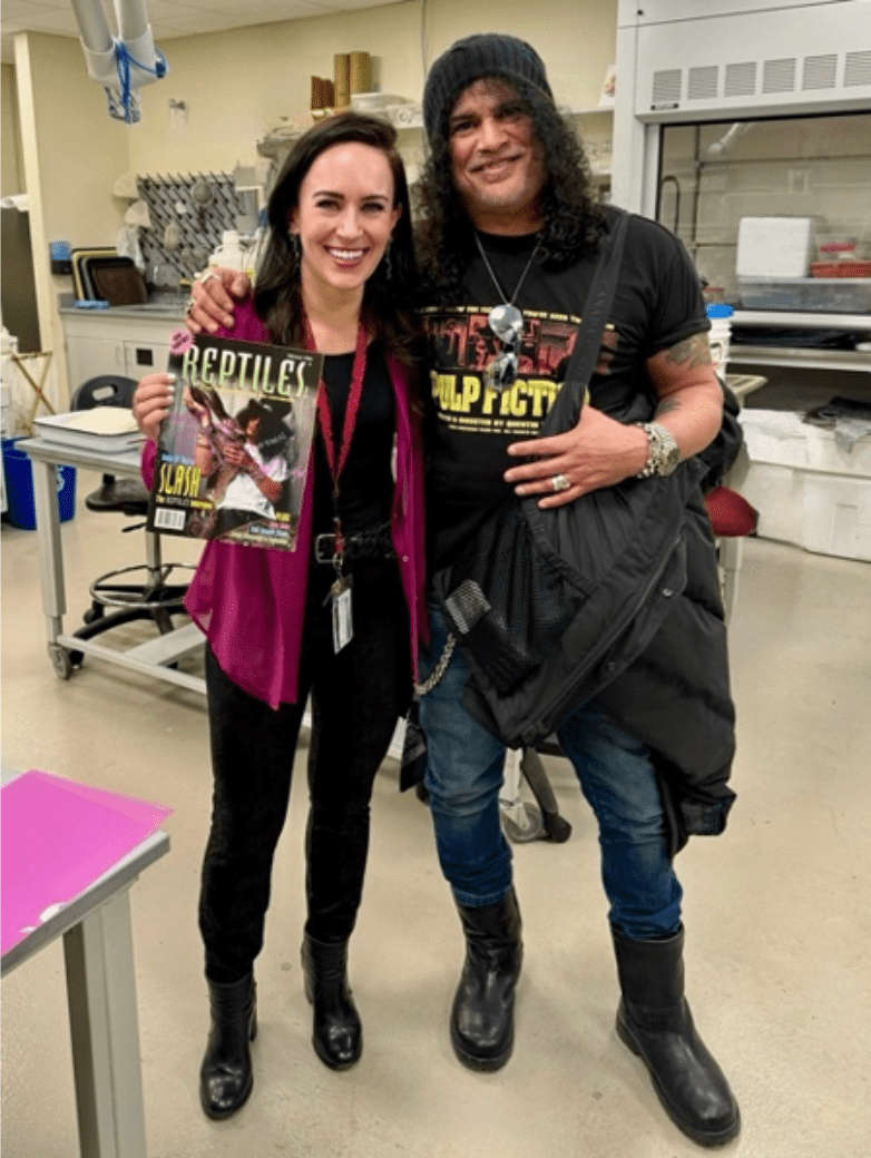 a scientists and a rock star stand in a lab with a copy of reptile's magazine. the rock star is on the cover of the magazine