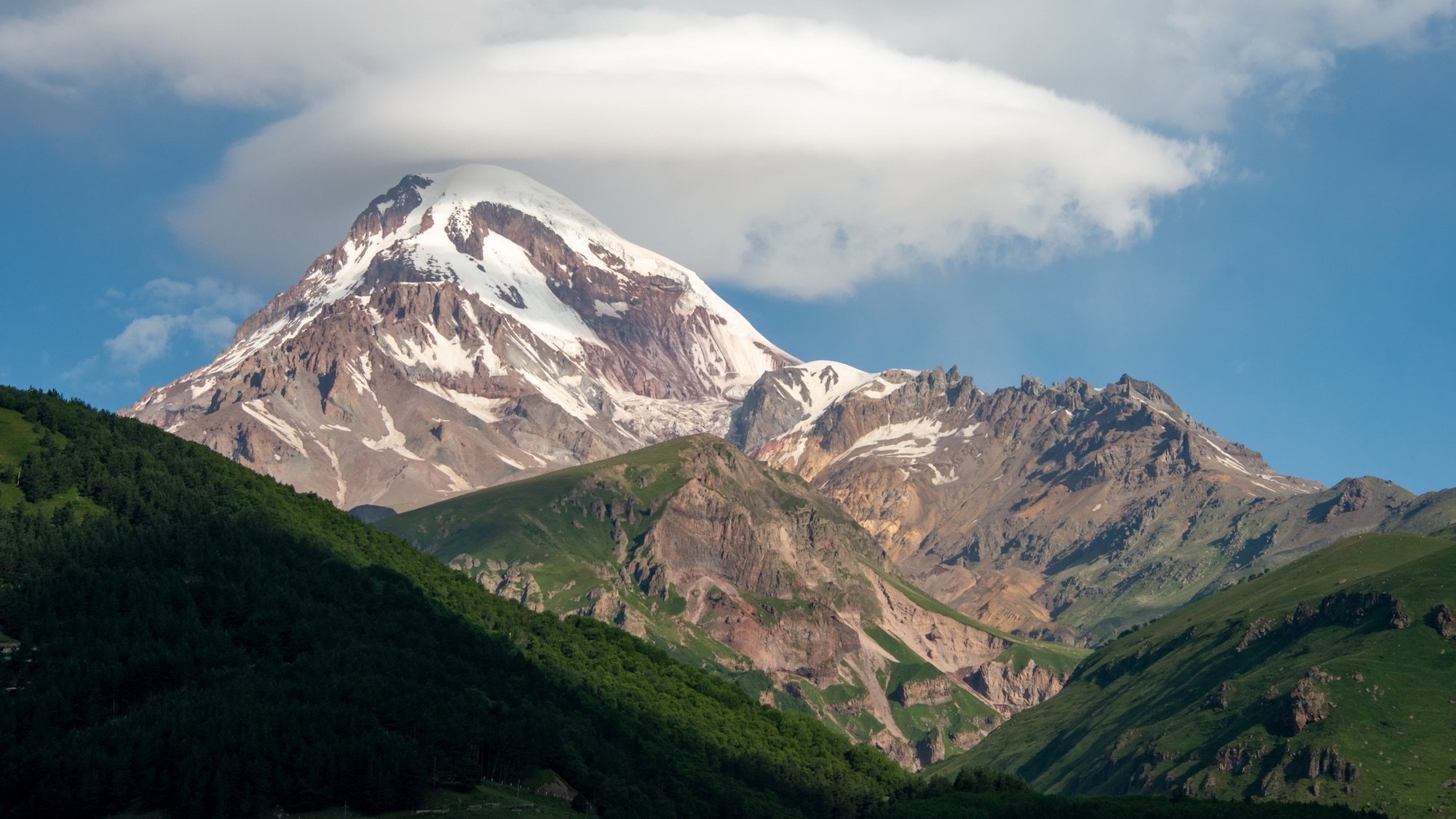 Snow-capped Mount Kazbek rising above lush green valleys and dramatic cloud formations in the Caucasus Mountains of Georgia, creating a powerful and iconic alpine landscape.
