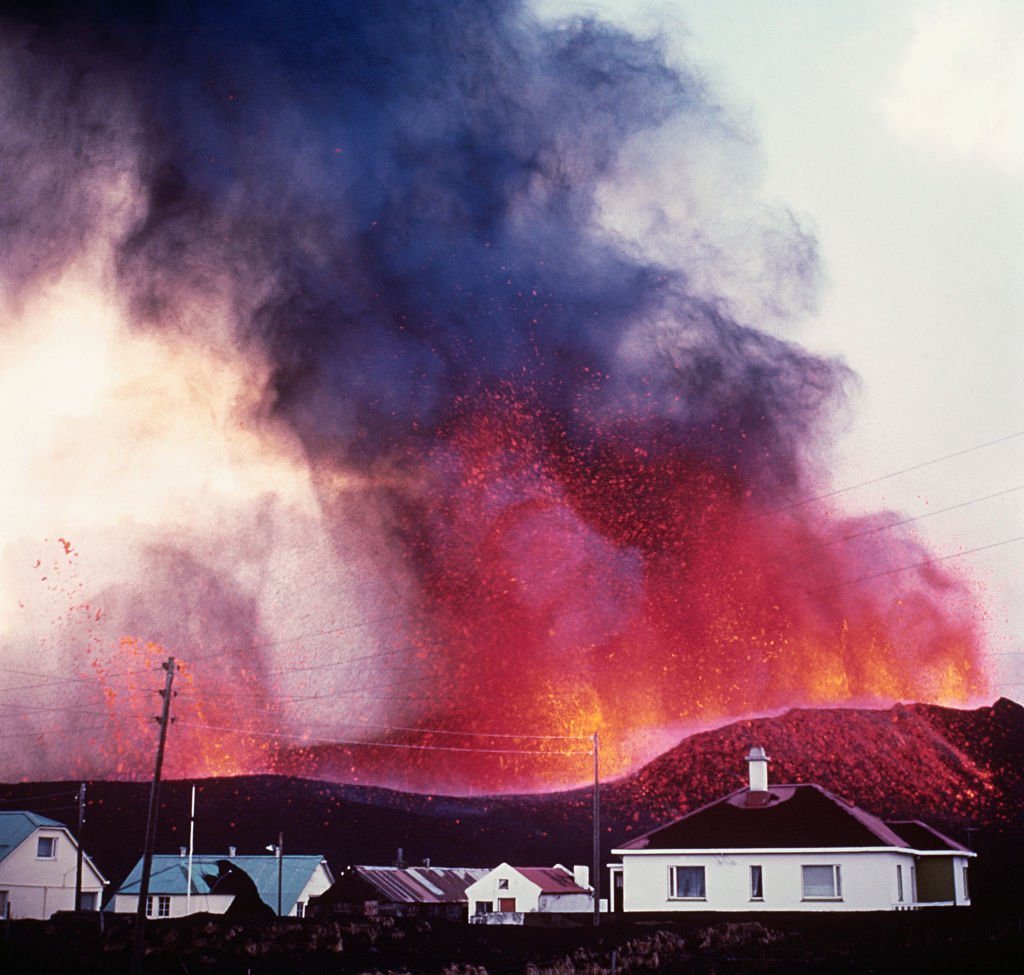 Vintage photograph showing several houses and telephone wires with volcano erupting in background.