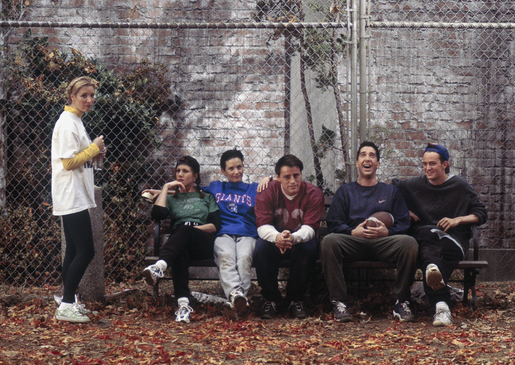 a still image from an episode of the show friends. a woman with blonde hair stands next to a bench while a woman with a black hat and green shirt, woman with a blue new york giants sweatshirt, man with a red jersey, man with a blue sweatshirt, and man with a black sweatshirt and blue hat on backwards sit on a bench above fall leaves