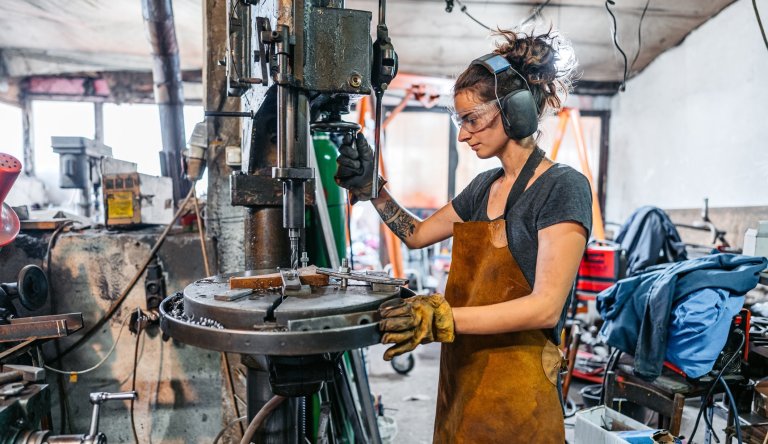 Young woman working on a drill press machine in a busy metal workshop.