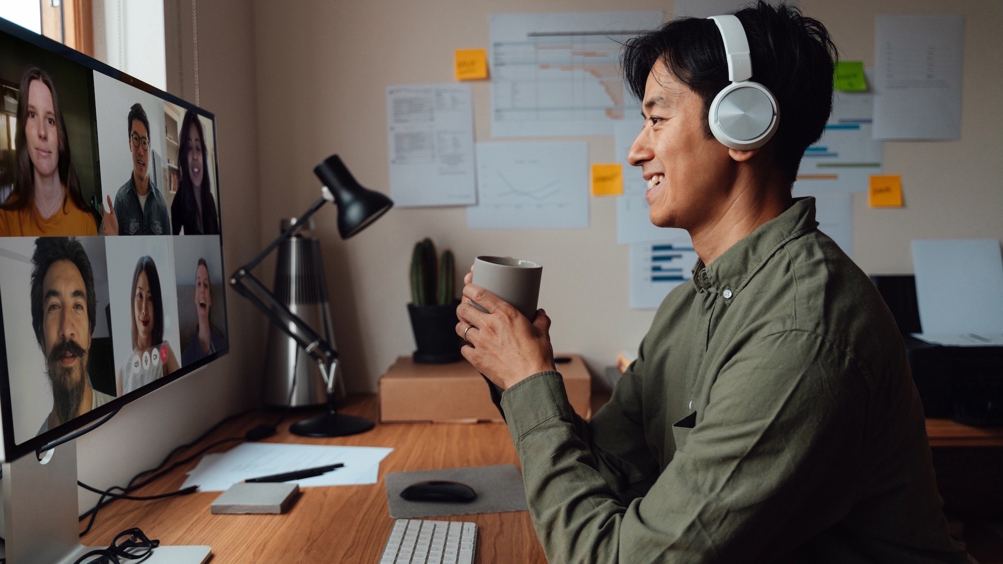Young man wearing wireless headphones, catching up with his team during a video call via desktop computer, sitting in his home office. Teleworking from home. Flexible working.