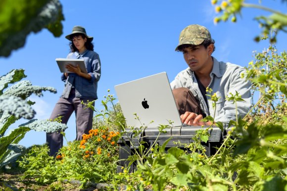 A man in a camouflage cap types on a silver 14-inch MacBook Pro set on a field case in a vegetable garden while a woman behind him takes notes on an iPad.