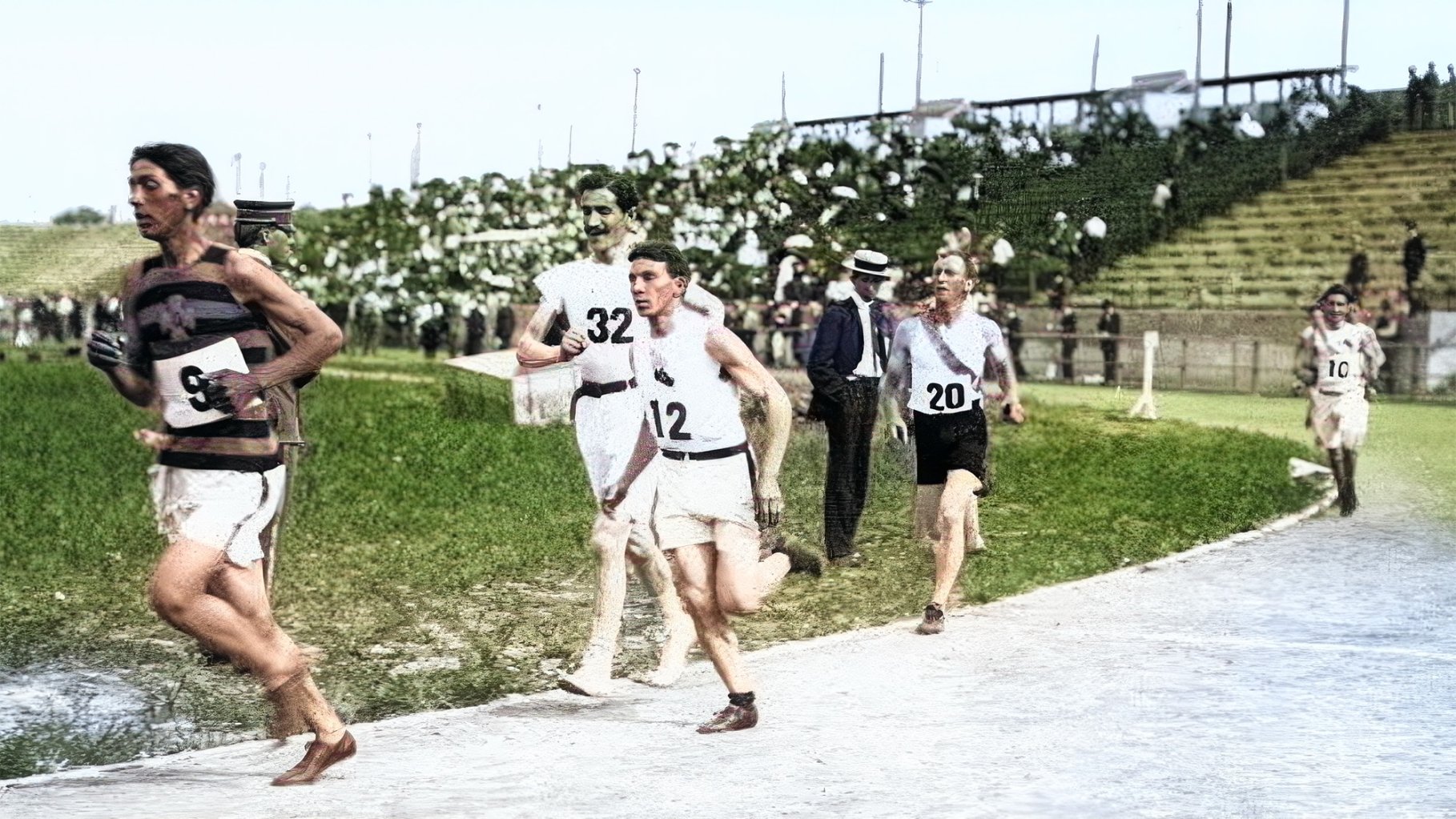 Vintage colorized photograph showing runners on a track with a crown of people in the stands in the distance.