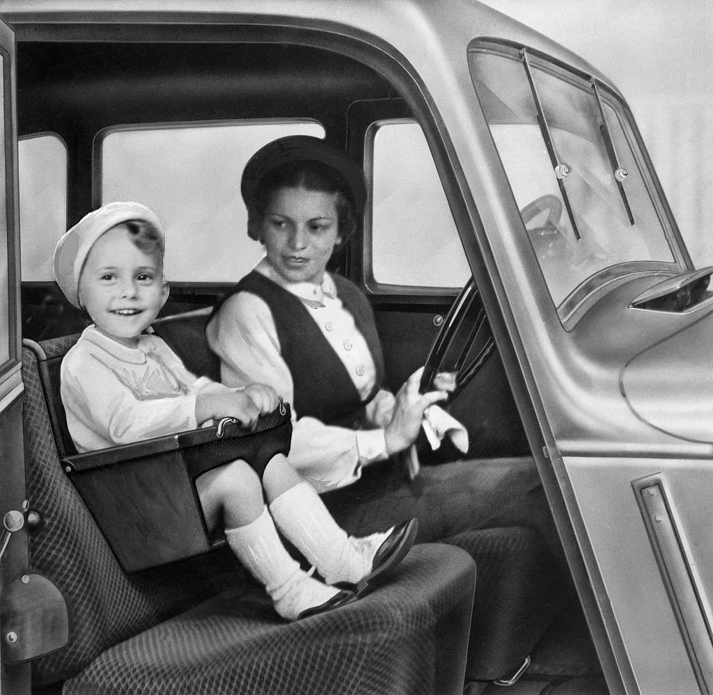 Small boy in a hat sits in a car seat next to a woman in vintage car. Photograph is black and white.