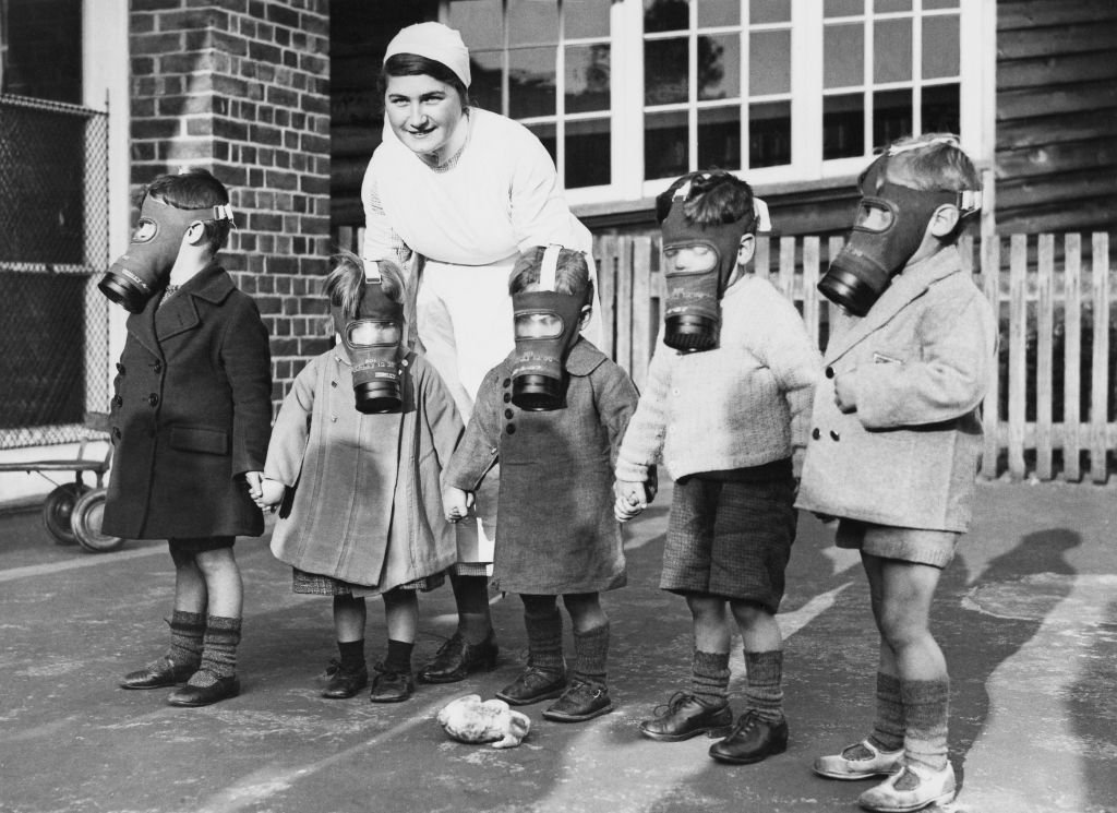 Nurse with five young children in gas masks in black and white photograph.