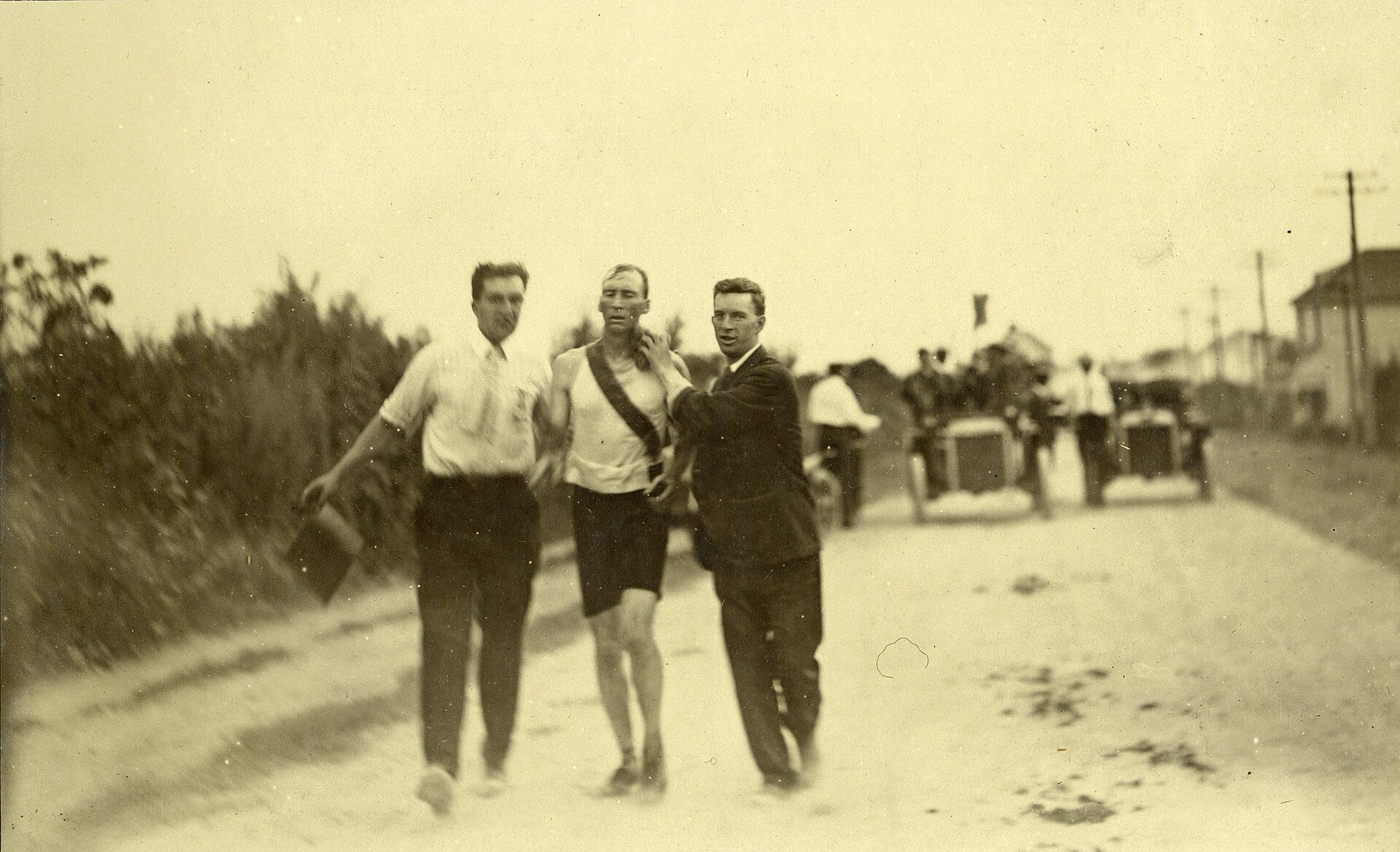 Historic photo showing two men next to a runner try to help him with vintage cars in the background.