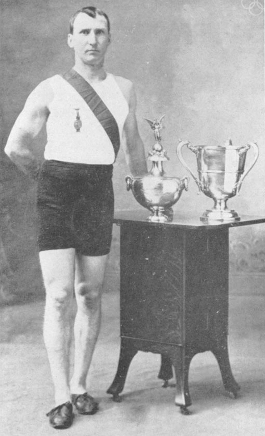Young white man with short hair stands with a sash next to a trophy.
