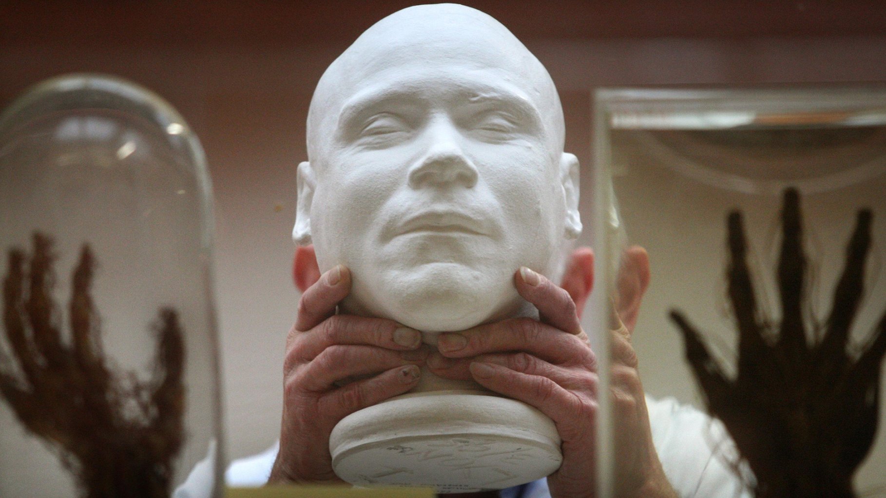Two hands holding up plaster cast of death mask surrounded by two jars with human hands.