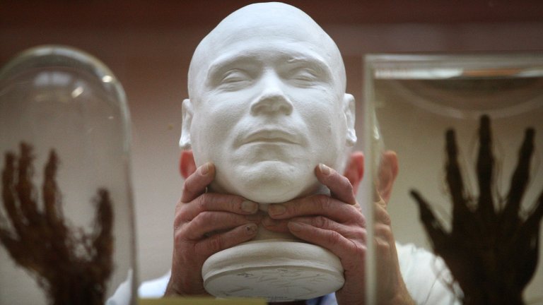 Two hands holding up plaster cast of death mask surrounded by two jars with human hands.