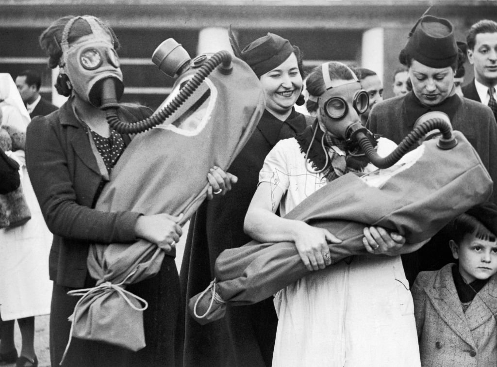 Two women hold baby-shaped infants in gas masks that completely surround the child.