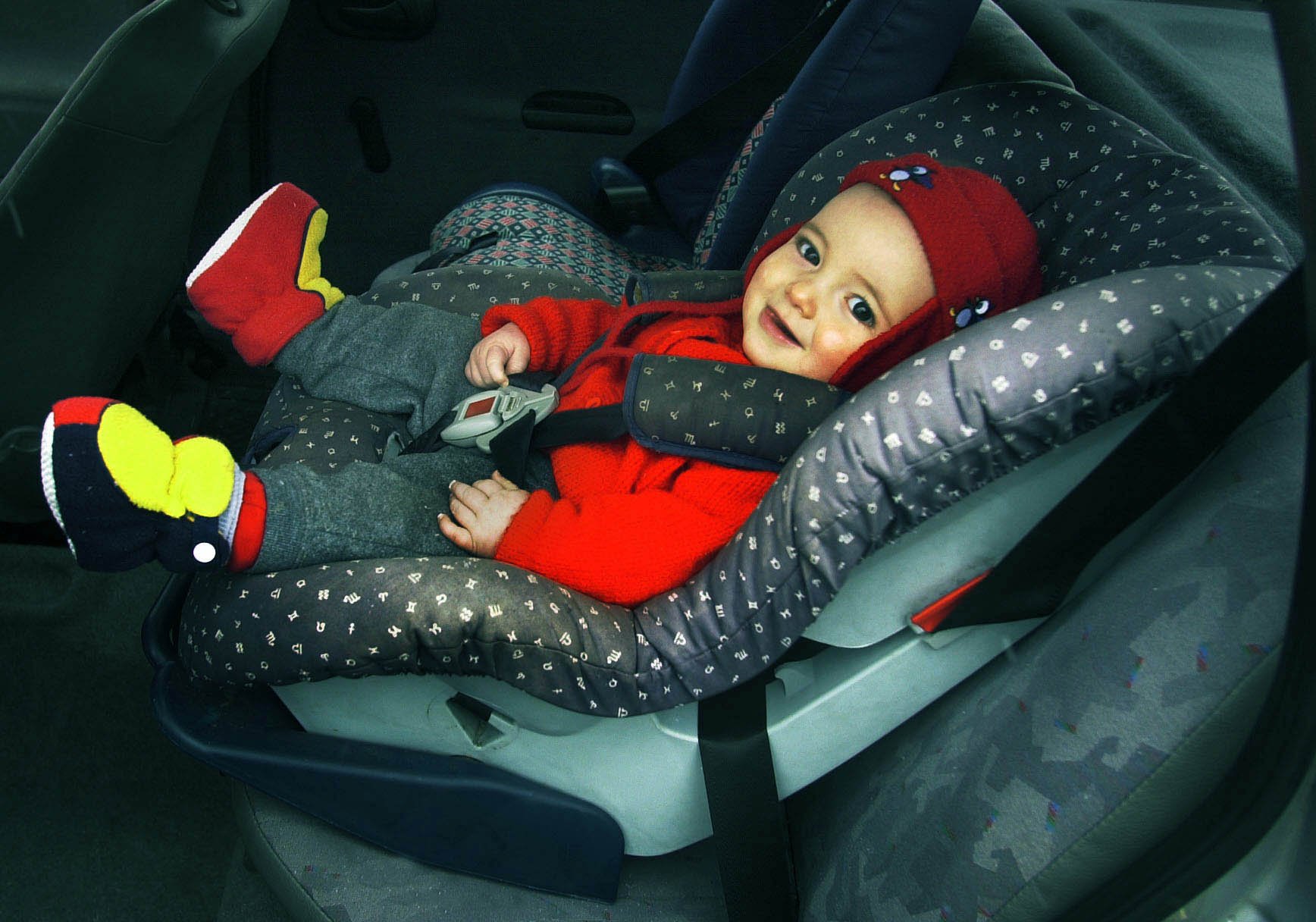 Baby in red hat and red and yellow shoes looks at camera from car seat.