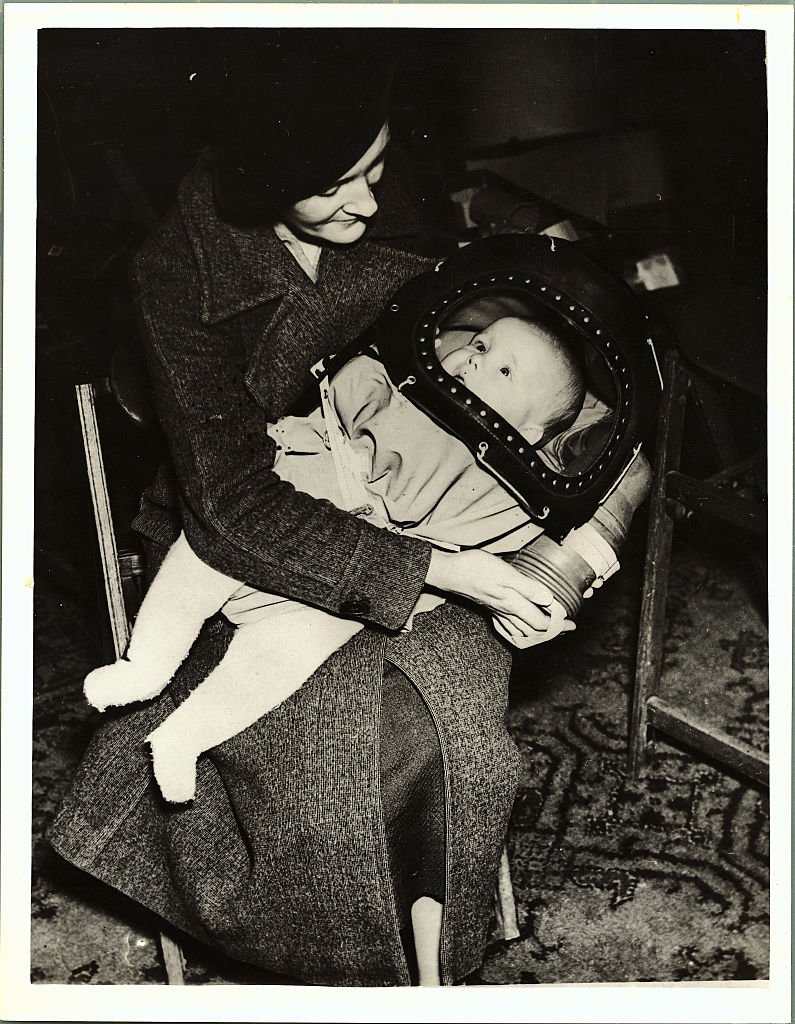Black and white photograph of woman in hat holding a baby in a hooded mask
