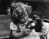 Little girl puts gas mask on doll in black and white vintage photograph.