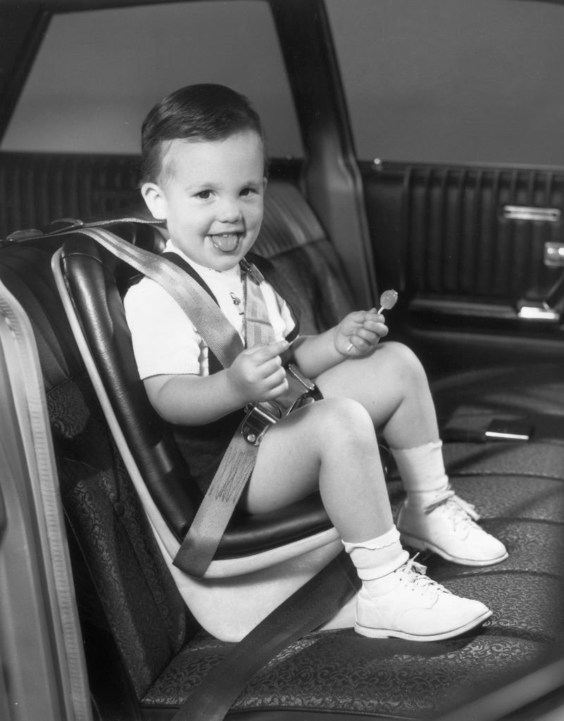 A small boy holding a lollipop sits in a carseat in a car, 1960s.