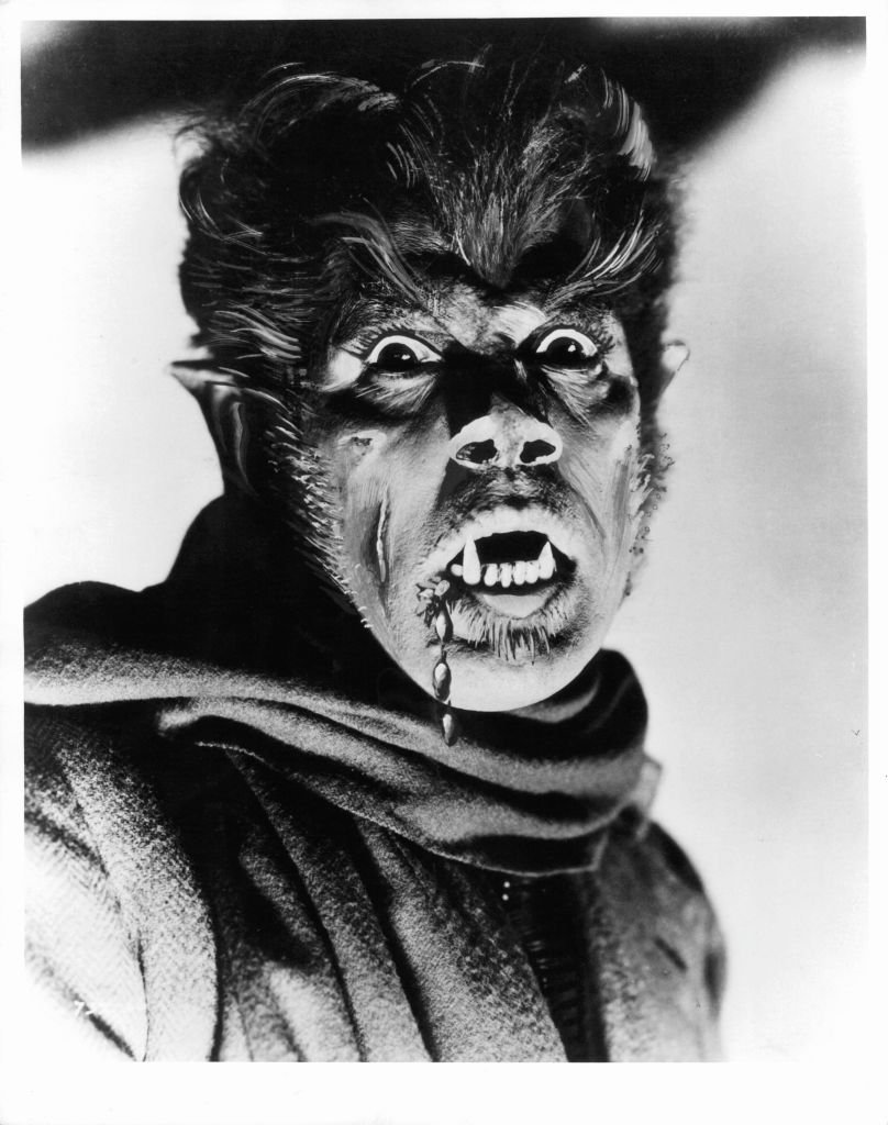 Headshot of actor in werewolf makeup with large pointed teeth and scarf.