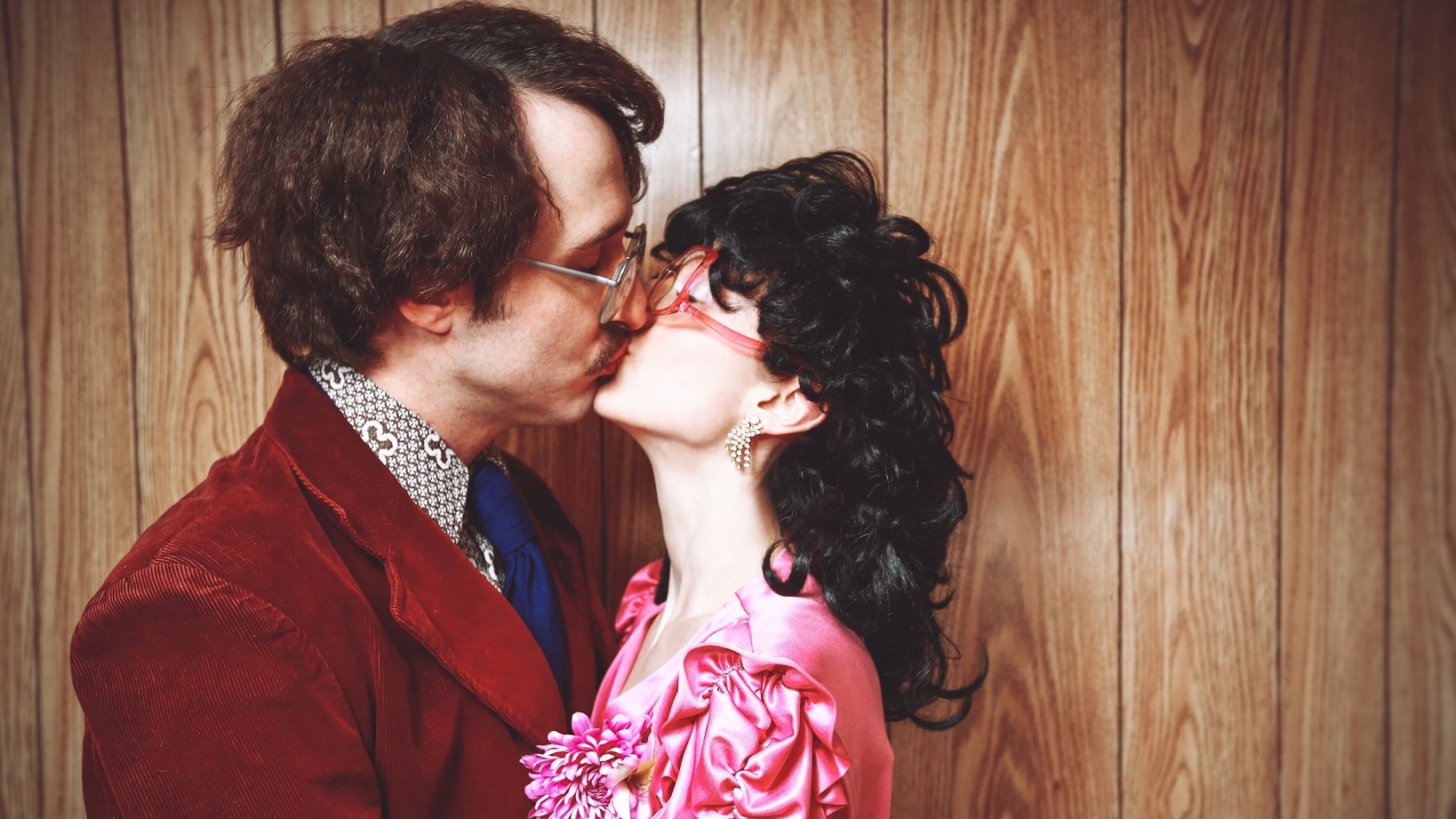 1970s couple kissing. Conceptual image for relationships, dating, and desire. The retro man is wearing a vintage suit and the retro woman is wearing a pink dress. Both have eyeglasses.